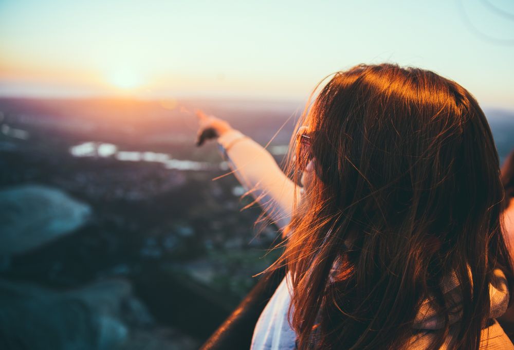 A woman points at the sun above a mountain.